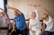 Four elderly people sat on chairs doing exercises with their arm reaching over their head